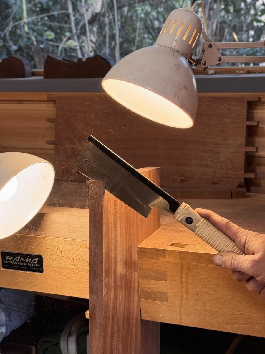 A dozuki saw cutting dovetails at the bench, under a lamp